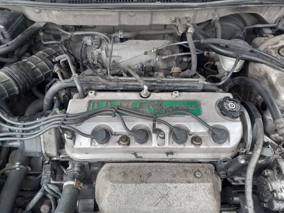 On the back of the valve cover is the PCV valve dead center and a vacuum hose on the left corner going to the air intake. You can also see the throttle linkage and cables behind the valve cover.