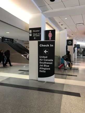 Signage right inside door from rental car shuttle near carousel #6 directing passengers to elevator for check-in for listed airlines. 