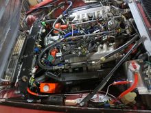 Cooling system overview. The alert will notice: a) no central spout on the crosspipe (a V12 saloon crosspipe is fitted which is the only new type now avaulable), b) the radiator purge tubes have gone, c) the radiator bleed is from the LHS - see red hose going to firewall header tank. The radiator filling bleed screw is in the banjo threads. The header tank is connected to the main system by a T into the heater return, the overflow is the red tank on the wing where the factory tank was.